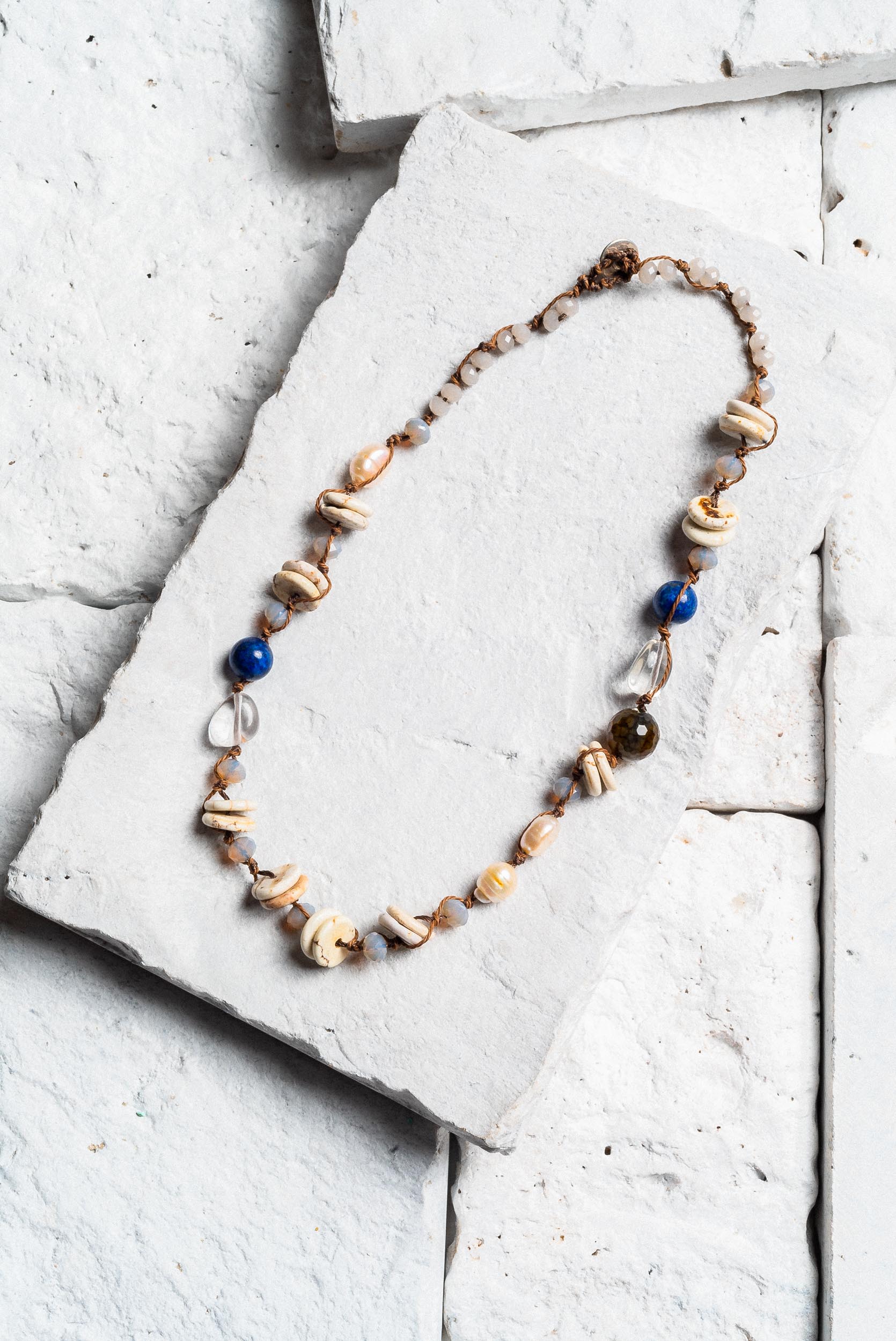 a choker necklace displayed on a white stone. The choker is made from brown leather strap with an even spread from end to end of shells and beads. There are clear white beads, small circular shells, large white beads, blue beads and dark beads as well as some pearls.